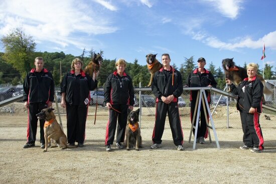 Teamfoto: geleiders in zwarte trainingspakken met hun Belgische Herders op het trainingsveld